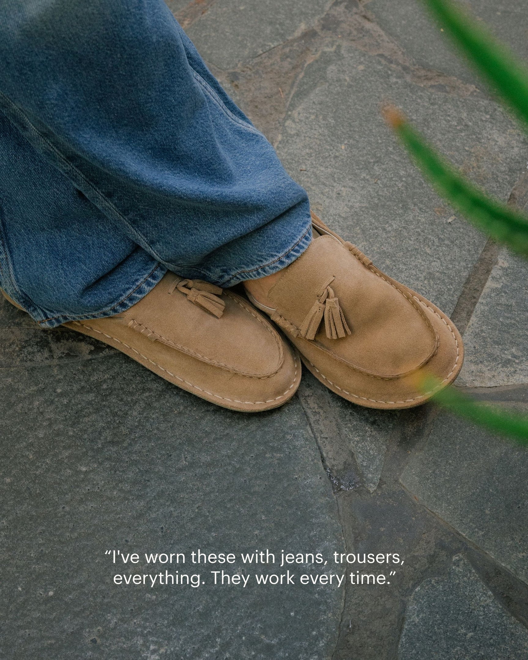 Tan suede loafers with tassel detail worn with blue jeans on stone pavement.