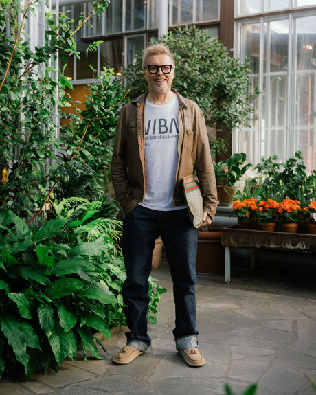 Man wearing tan suede desert boots with dark jeans, light blue graphic t-shirt, and brown cardigan in a greenhouse setting.