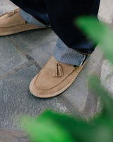 Tan suede loafer with tassel detail worn with jeans on concrete surface.